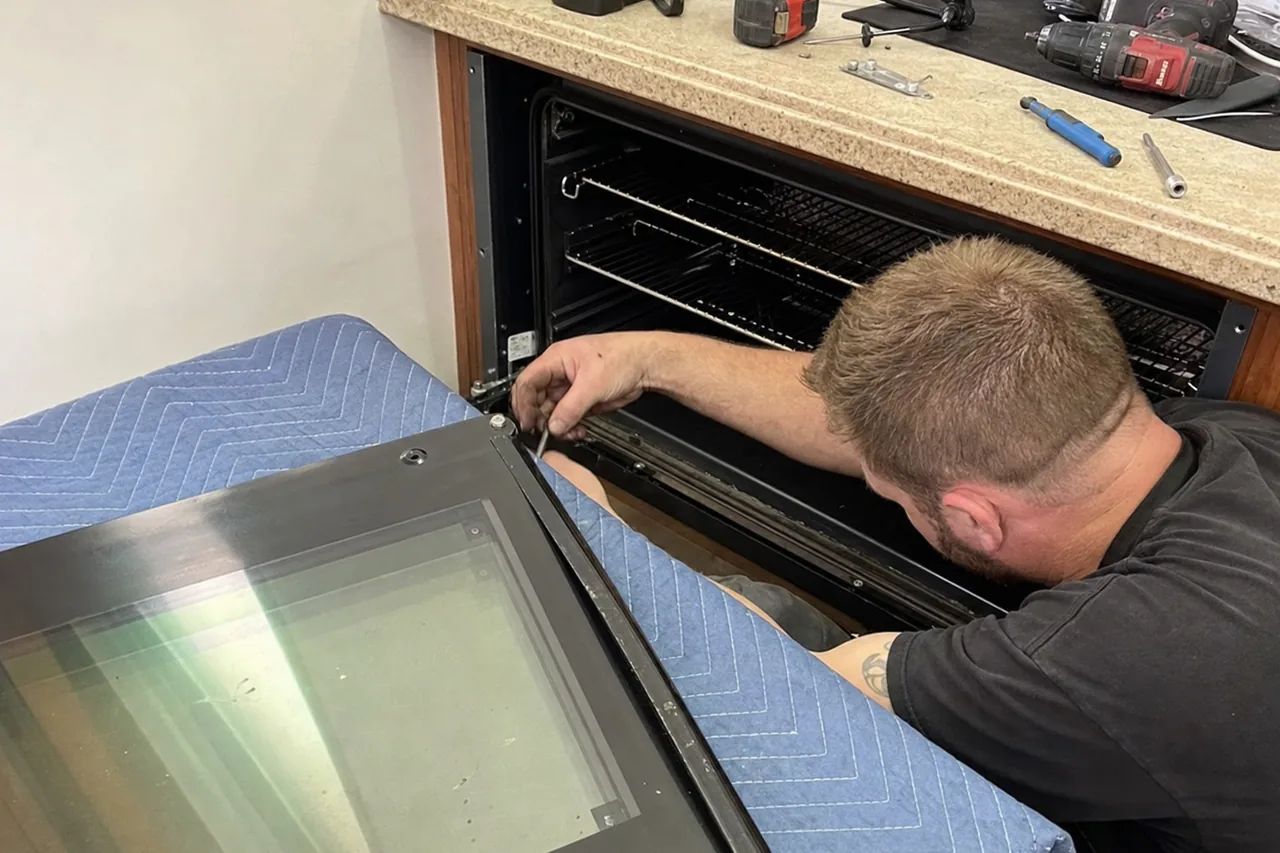 A technician kneeling at an open wall-oven cavity with his hands at the hinge receivers and tools laid out on the counter above