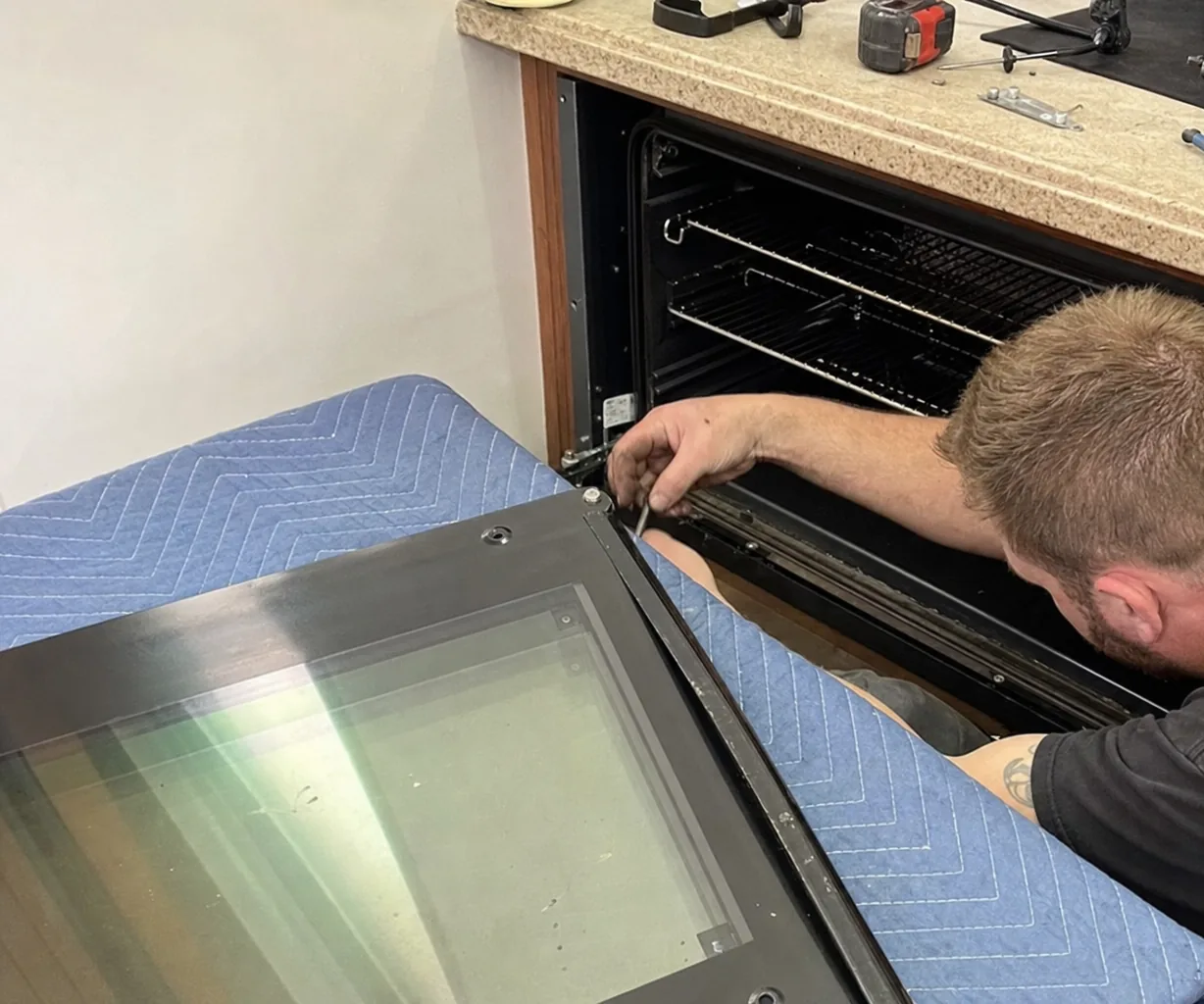 Left-side view of a removed oven door on a quilted blanket beside the open appliance cabinet
