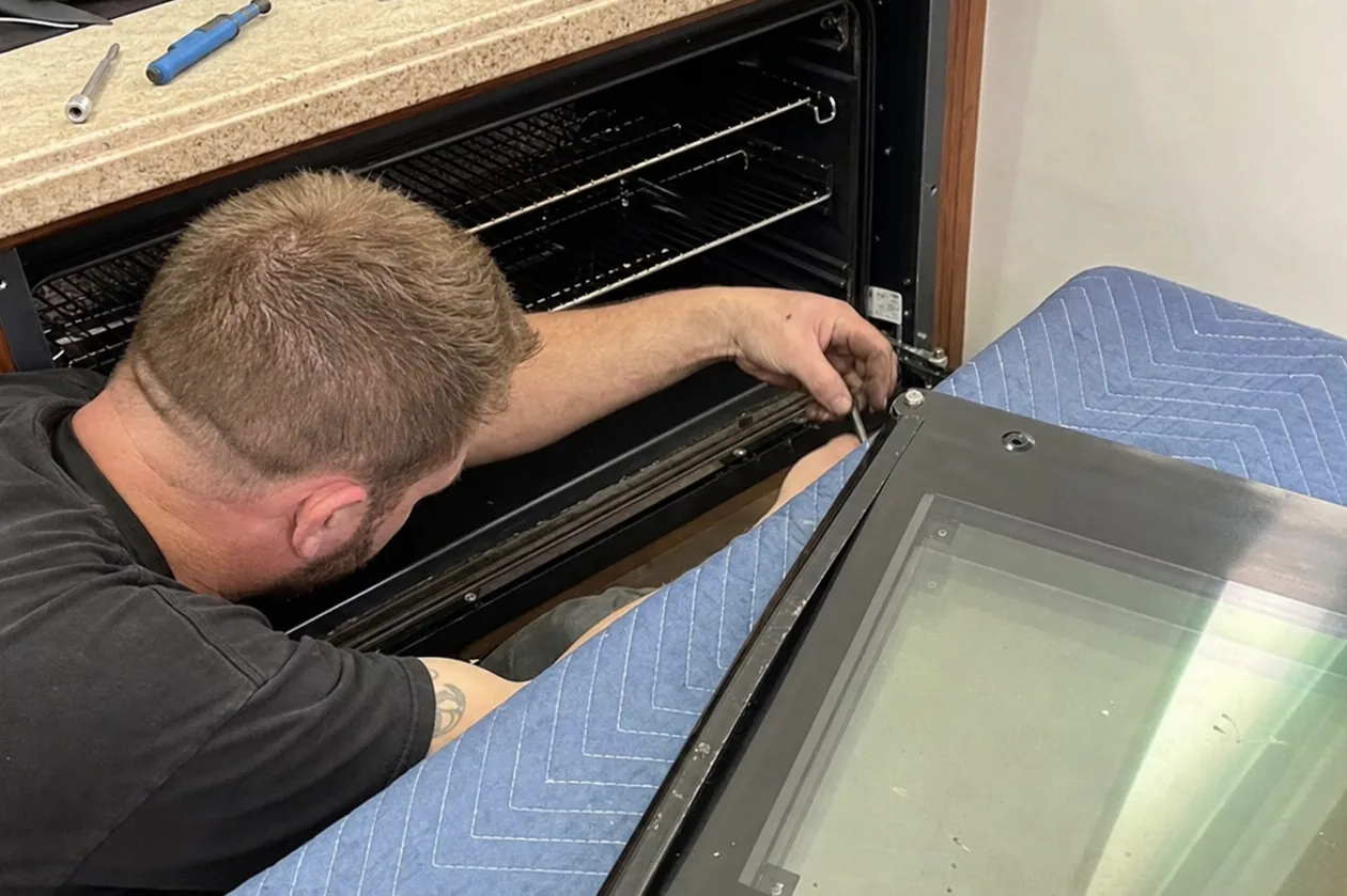 Angled crop of a technician working inside a wall-oven cabinet with the door panel on a blanket at the left edge