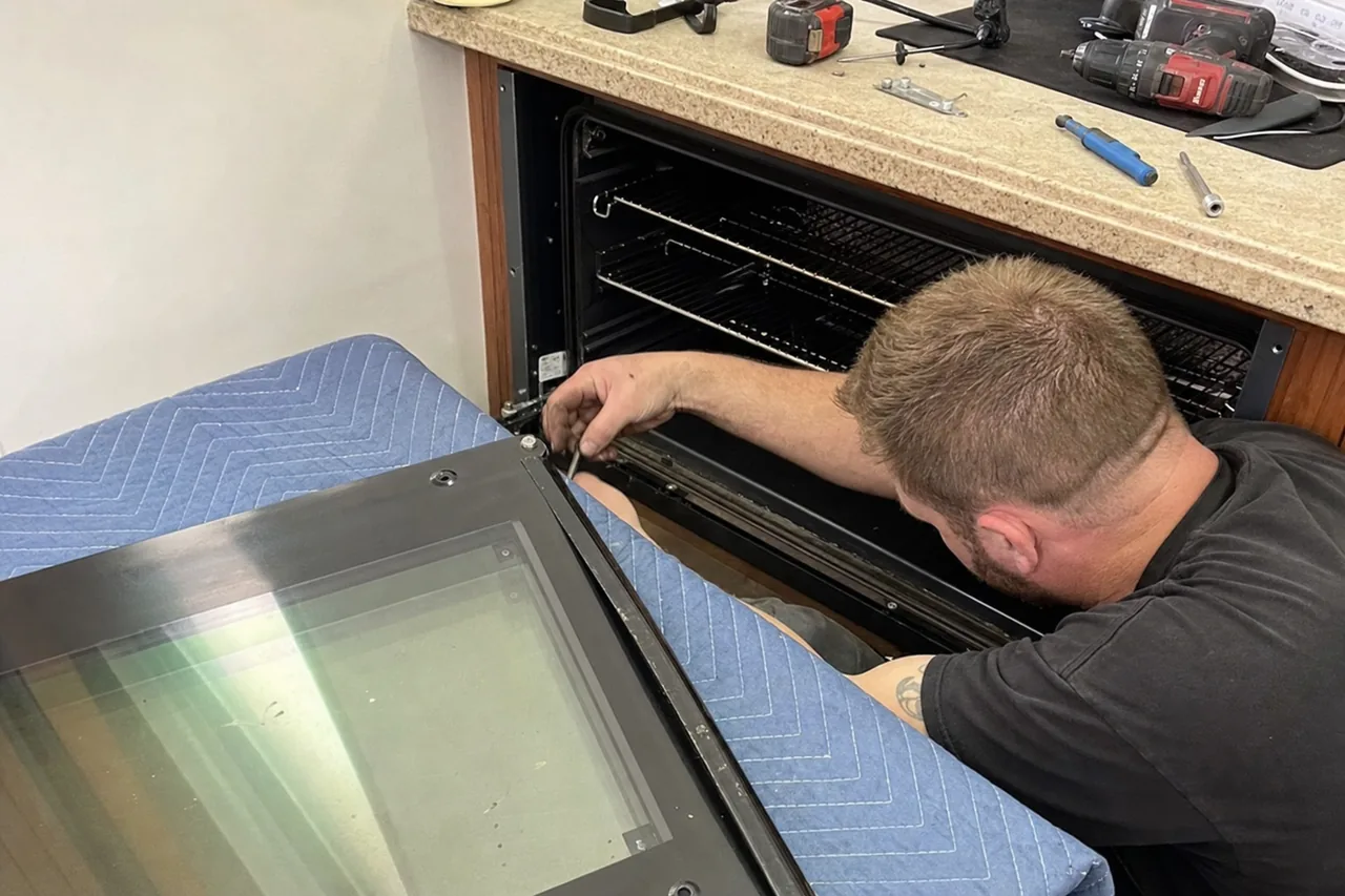 A built-in wall oven with its door removed and laid face-up on a padded blanket while a technician works at the exposed hinge slots